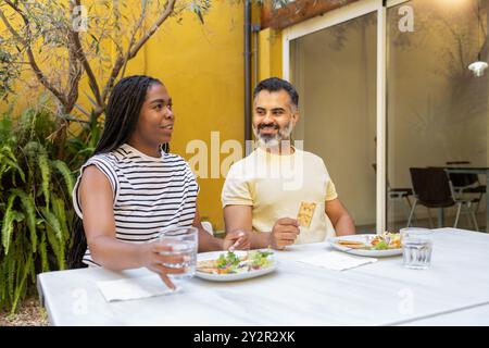 A cheerful black woman and an indian man indulge in a pleasant conversation while savoring a meal outdoors The vibrant setting adds a soothing backdro Stock Photo
