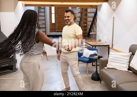A black woman and an indian man happily dance together in a stylish living room, showcasing joy in their comfortable home environment Stock Photo