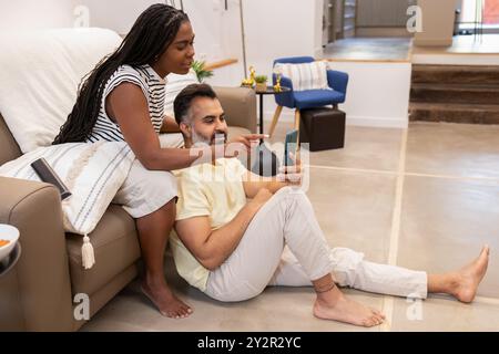 In a modern living room, a black woman shares something on phone with an indian man seated on the floor, both appearing relaxed and comfortable Stock Photo
