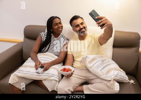 A happy black woman and an indian man seated on a sofa, smiling as the man takes a selfie The warm indoor setting highlights a shared relaxed moment w Stock Photo