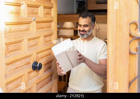 A cheerful middle-aged indian man receives a package at his home's wooden front door, expressing happiness Stock Photo