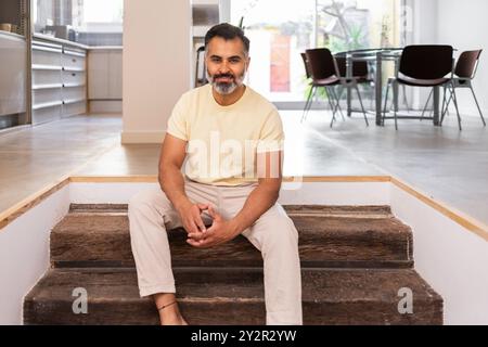 A middle-aged indian man enjoys a quiet moment sitting on wooden stairs in a well-lit home interior, reflecting a comfortable, serene atmosphere Stock Photo