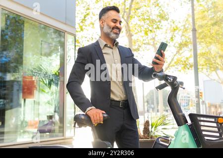 A cheerful Indian man in business attire interacts with his smartphone while leaning on an electric bike in an urban setting, showcasing a blend of mo Stock Photo