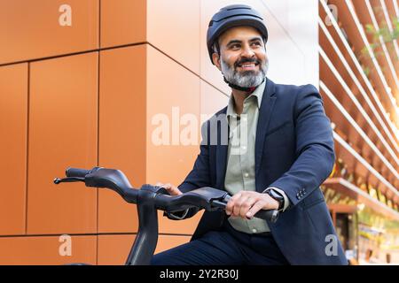 Smiling Indian man in formal attire rides an electric bike in a city, looking away from the camera. Stock Photo