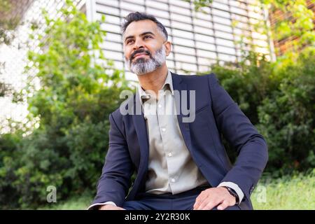 From below an Indian man in a dark suit seated outdoors, gazing away with a thoughtful expression, in a lush urban setting. Stock Photo