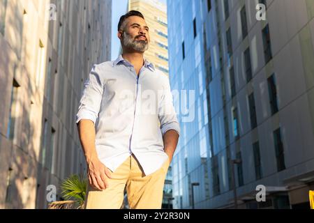 From below a stylish Indian businessman in a light blue shirt and khaki pants looks thoughtfully away amidst urban surroundings. Stock Photo