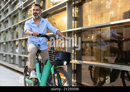 Middle-aged Indian man cycling on an electric bike through a city street, casually dressed and looking away from the camera. Stock Photo