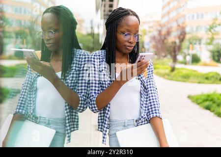 A focused black woman manages her busy schedule by using a smartphone and carrying a laptop in an urban business district Stock Photo