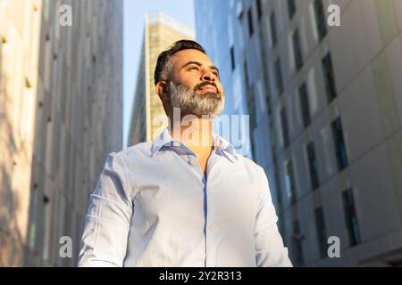 From below an Indian man dressed in formal attire, looking away thoughtfully, against an urban backdrop with modern architecture. Stock Photo