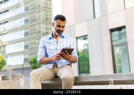 A mature Indian man, dressed in a formal light blue shirt and beige trousers, uses a tablet while sitting outdoors on a bench in an urban setting. Stock Photo