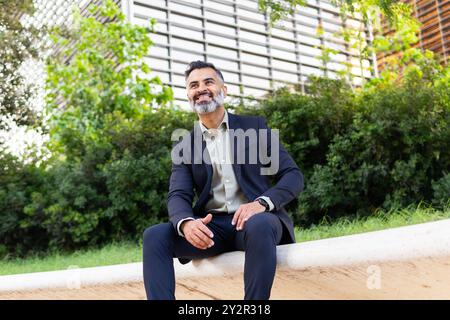 From below man in formal business attire is seated outdoors with a modern building in the background. He looks away from the camera, presenting a rela Stock Photo