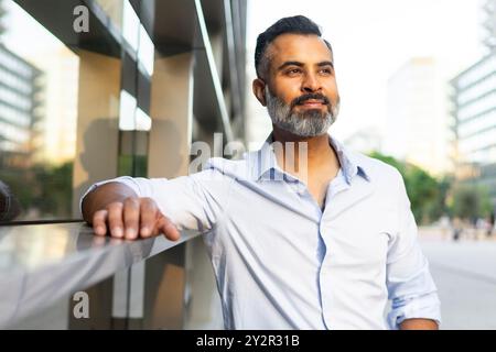 An Indian businessman with a stylish beard, wearing a light blue shirt, looks thoughtfully away from the camera while leaning against a modern glass r Stock Photo