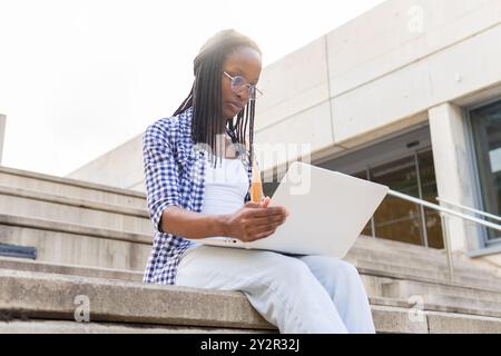 A Black woman, business casual in a plaid shirt and sunglasses, is engaged with her laptop and documents while seated outdoor on steps in a business d Stock Photo
