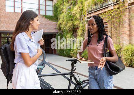 Two multiethnic college students engaging in a lively discussion while standing next to a bicycle on a university campus, surrounded by lush greenery Stock Photo