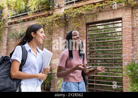 Two cheerful multiethnic students chatting and laughing as they walk through a lush university campus, carrying books and a laptop Stock Photo