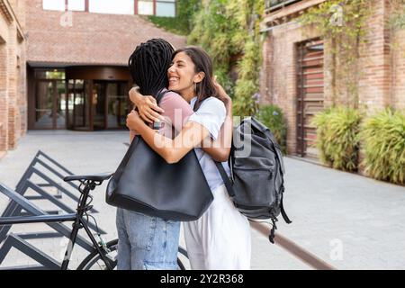 Two female students embrace joyfully in a university campus courtyard, happy to reunite, carrying backpacks Stock Photo