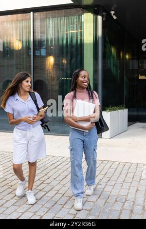 Two young female college students walk together on a university campus, engaging in conversation with books and a laptop in hand, clad in casual attir Stock Photo