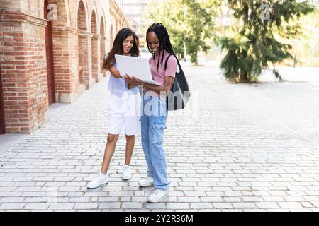 Two diverse female students joyfully engage with a laptop outside a brick university building, pointing at the screen and discussing academic content Stock Photo
