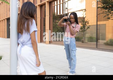 A young, cheerful female student captures photos of her stylish friend outside a modern university building Stock Photo