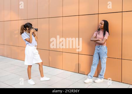 A young Asian woman uses a camera to take a photo of her African friend who poses confidently with arms crossed against a uniform orange wall Stock Photo