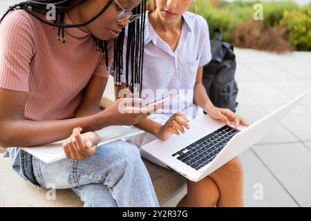 Two college students collaborate on their studies using a laptop and smartphone while seated outside on their university campus Stock Photo