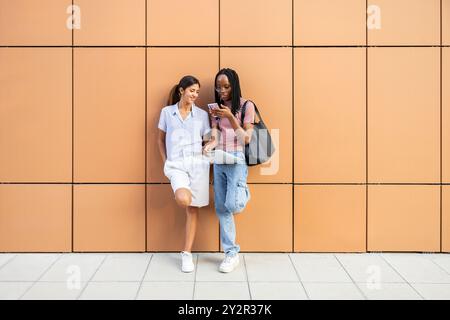 Two young female students share a moment while using a smartphone against a modern orange wall background They have casual style and engaging in a che Stock Photo