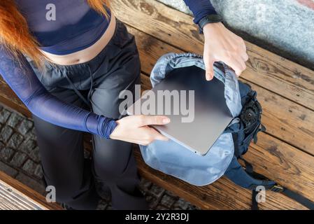A modern young woman with orange hair is observed inserting a slim laptop into her urban-styled backpack She is dressed in contemporary athletic attir Stock Photo