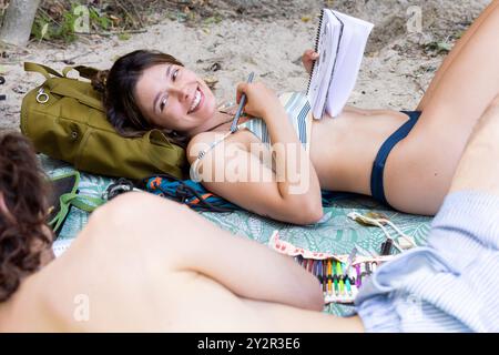 A young woman smiles joyfully while lounging by the River Alberche, accompanied by friends on a sand riverside, surrounded by nature and items for a d Stock Photo