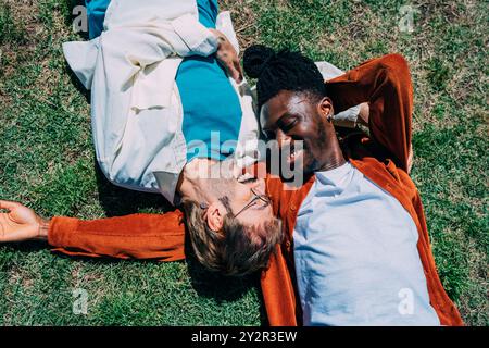 A joyful moment shared between a gay couple, with an African American man and his partner laughing while lying on the grass in a sunny city park, show Stock Photo