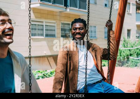 Multiethnic gay couple smiling and enjoying time together on a playground swing in the city, reflecting happiness and love Stock Photo