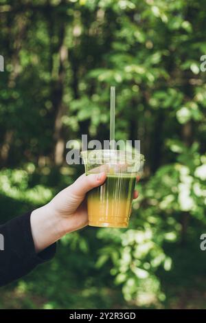 Cropped unrecognizable teenager girl holding a refreshing iced matcha drink in a clear plastic cup with a straw against a lush green foliage backdrop Stock Photo