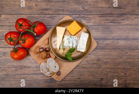 Top view of a wooden plate with various cheeses, fresh basil, and ripe tomatoes alongside bread and nuts on a rustic wood background, portraying a hea Stock Photo