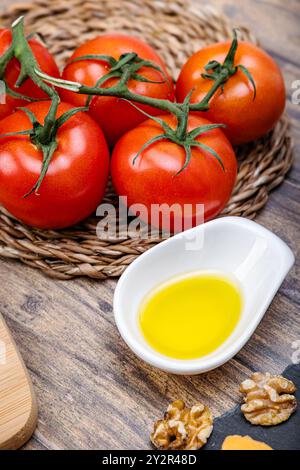 Vibrant red tomatoes on a vine and a small white bowl of olive oil, arranged on a rustic wooden table with walnuts and a wicker basket. Stock Photo