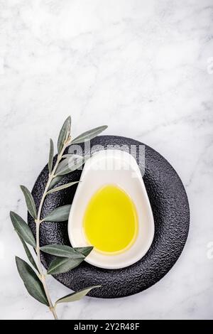 Top view of a simple, elegant presentation of olive oil in a white ceramic dish, set on a textured black plate surrounded by fresh olive leaves on a m Stock Photo
