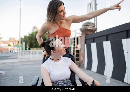 A joyful moment between two women, as one points out something interesting to her friend in a wheelchair They are outdoors, expressing happiness and c Stock Photo