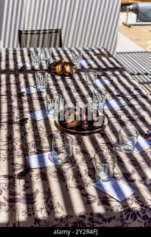 An outdoor dining table is bathed in sunlight, casting striking shadows across a patterned tablecloth. Slices of bread and beautifully-prepared food a Stock Photo