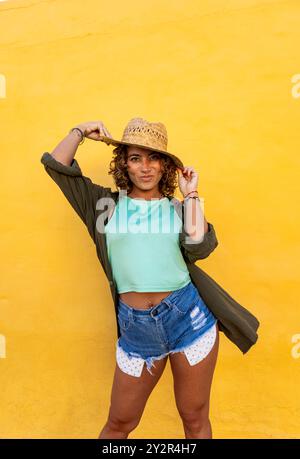 A woman with curly hair playfully adjusts her straw hat against a vibrant yellow background. She is standing confidently, dressed in a casual summer o Stock Photo