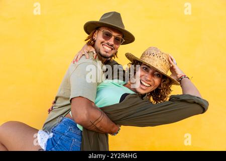 A cheerful man with dreadlocks and a smiling woman with curly hair have fun in a playful pose against a bright yellow background. Both are wearing hat Stock Photo