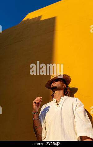 A man with dreadlocks looking away from the camera, wearing a straw hat and a white shirt, stands in contrast to a vibrant yellow background. Stock Photo