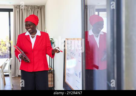 A professional African modern businesswoman in a vibrant red suit and matching headscarf smiles while engaging in a presentation, pointing at a whiteb Stock Photo