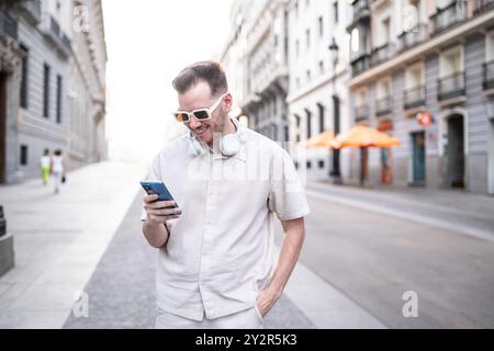 A trendy young man with headphones around his neck smiles as he uses his smartphone on a sunny, urban street. Stock Photo