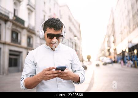 A cheerful young latin man enjoys his music using headphones while managing apps on his smartphone, walking through a sunlit urban street. Stock Photo