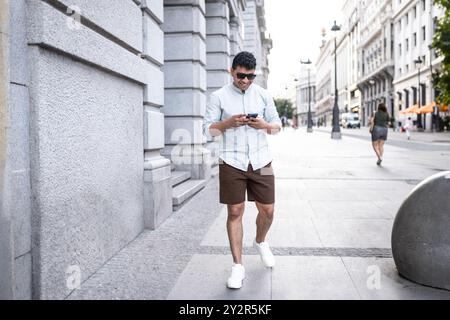 A young latin man dressed in a casual white shirt and brown shorts smiles as he uses his smartphone while walking on a bustling city street. Stock Photo