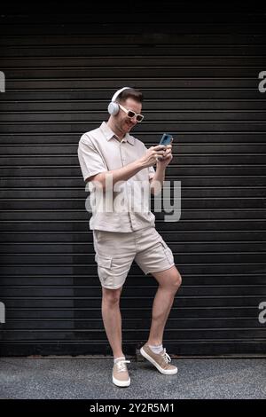 A cheerful young man wearing sunglasses and headphones stands against a dark textured wall. He is using a smartphone, smiling while choosing songs or Stock Photo