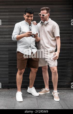 A joyful multiethnic gay couple stands against a striped dark backdrop, sharing a laugh while looking at a vintage camera together. The image captures Stock Photo