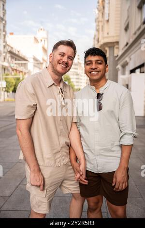 A cheerful multiethnic gay couple standing together on a city street, smiling at the camera while holding hands, showcasing a candid moment of love an Stock Photo