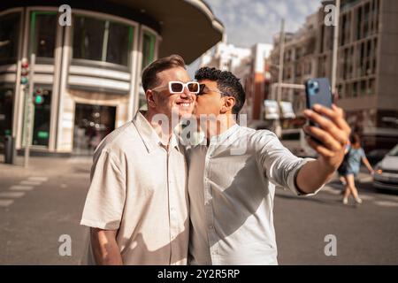 A joyful multiethnic gay couple captures a kiss and a selfie moment together, amidst the bustling streets of a sunny urban environment. Stock Photo