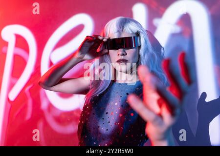 A futuristic woman, with long white hair and visor glasses, stands in front of a digital backdrop. Her attire glows subtly, showcasing a tactile conne Stock Photo