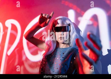A young woman with silver hair and futuristic glasses stands against a vibrant red backdrop, actively interacting with a cutting-edge digital interfac Stock Photo