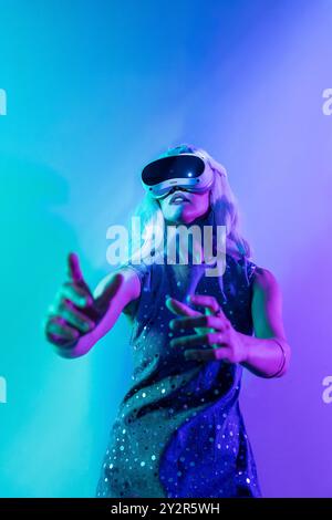 A woman engages with a virtual reality headset under vibrant blue and purple lights, gesturing with her hands as if interacting with a digital world. Stock Photo